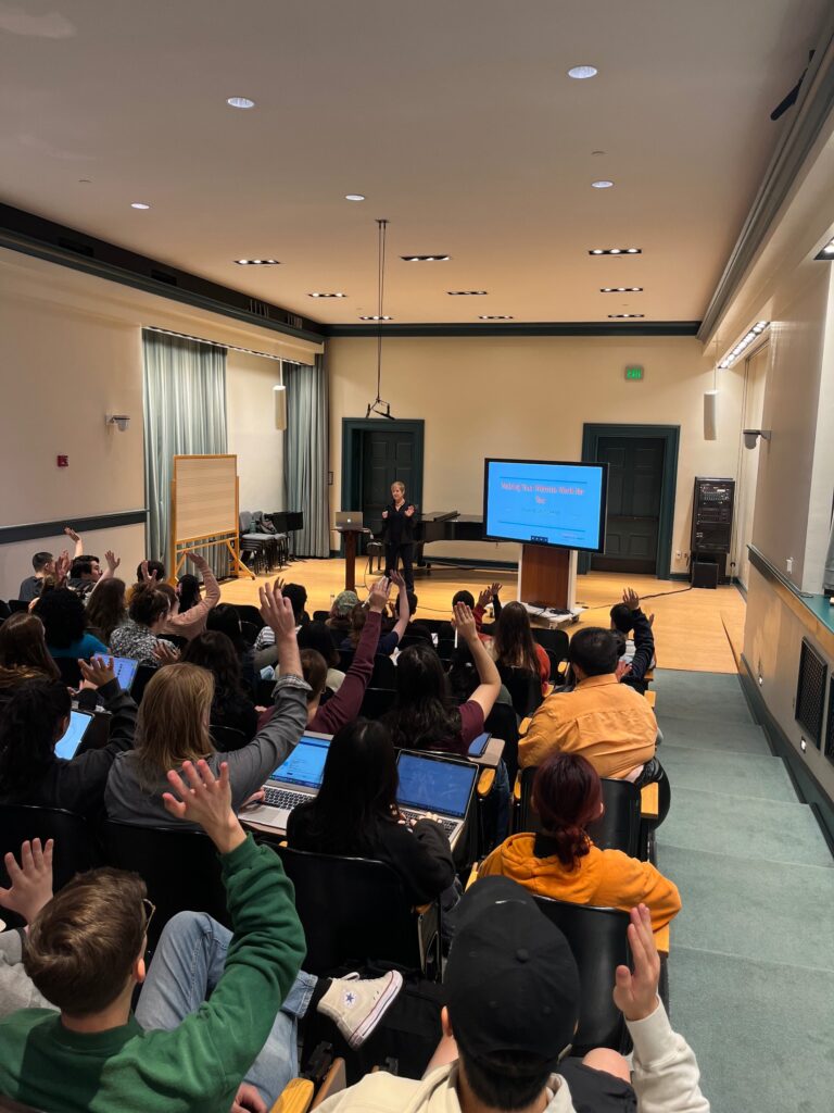 Composer Alexandra Gardner presenting on a stage to a group of students in an auditorium.